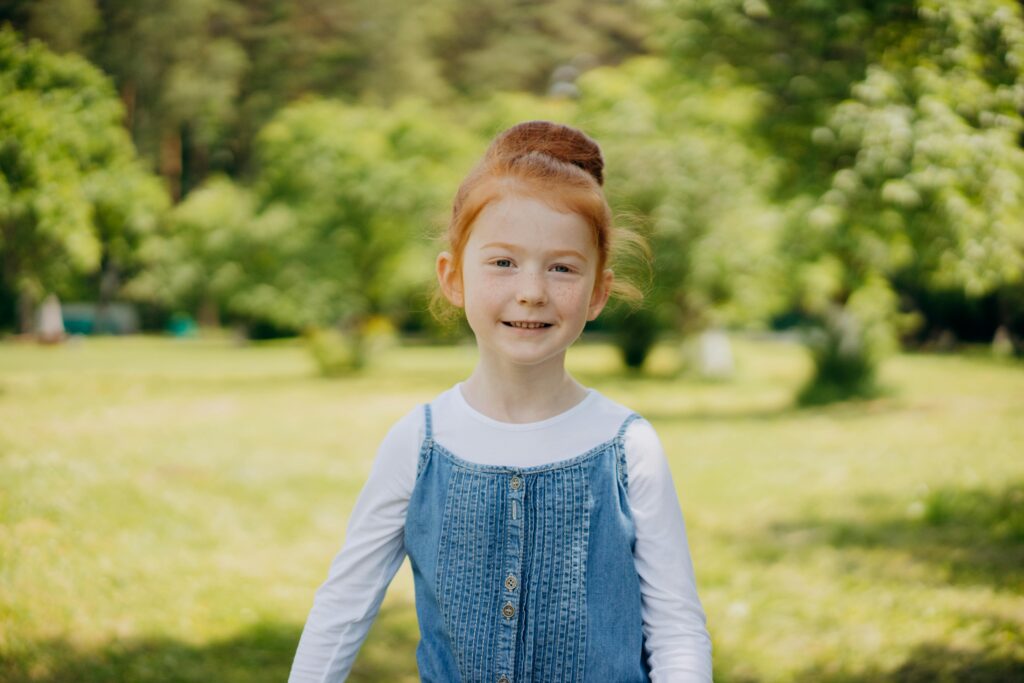 A cheerful redheaded girl smiling in a sunny, green park setting, enjoying a beautiful day outdoors.